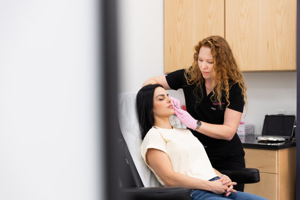 A patient at Horizon OBGYN & Aesthetics sits on a treatment chair as their provider carefully places their cheek filler in Winter Garden. The provider, wearing scrubs and surgical gloves, administers the filler in specific areas of the cheeks to restore volume.